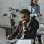 Pensive man using laptop in modern office with female colleague in background.